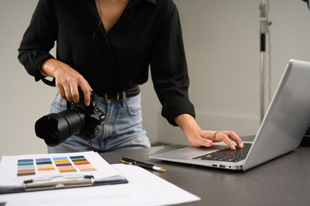 Cropped shot female photographer holding digital camera and using laptop at studio.