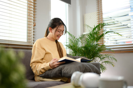 Smiling teenage woman wearing casual clothes reading book on couch at home.の写真素材