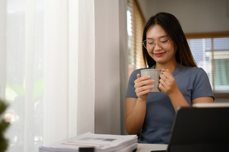 Happy young Asian female freelancer enjoying her cup of coffee while working at home office.の写真素材