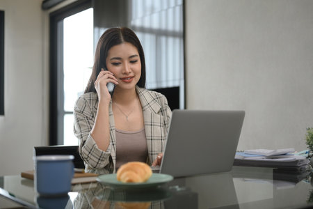 Beautiful young woman talking on mobile phone and working with laptop at home.の写真素材