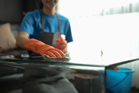 Cleaning service worker in wearing rubber gloves wiping glass table with cloth in modern home.の写真素材
