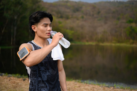 Athletic man standing near lake and drinking water after workout outdoors.