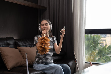Carefree young woman in headphones cleaning house and singing, using duster as microphone.