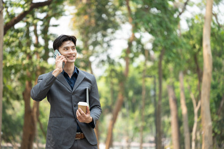 Confident Asian businessman with takeaway coffee talking on mobile phone outside on a sunny day.の写真素材