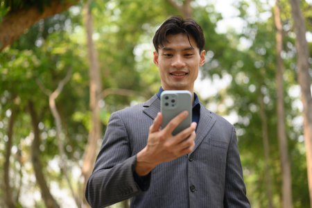 Confident young businessman text messaging on smart phone standing in city park.の写真素材