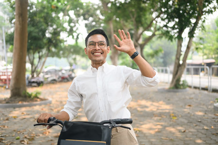 Cheerful asian businessman walking with his bicycle and waving hand for greetings, saying hello.の写真素材