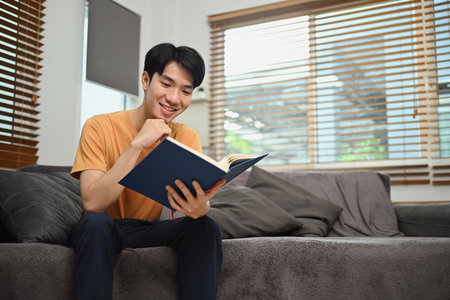Handsome asian man reading book on couch at home. Hobby, leisure and knowledge concept.の写真素材