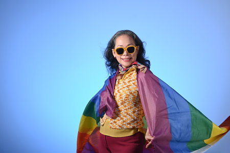 Confident middle woman wearing sunglasses wrapped in a rainbow flag on blue background. LGBT...