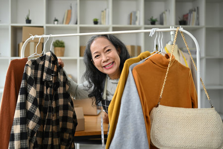 Middle age asian woman small business owner standing near clothing rack and smiling cheerfully.の写真素材