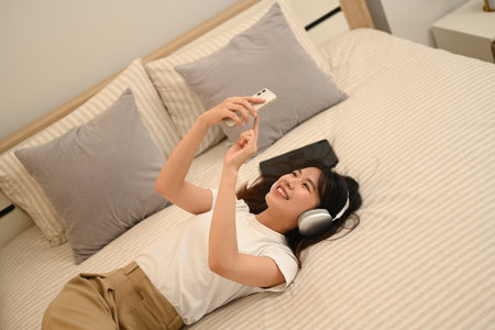 Smiling young woman listening to music with headphones and using smartphone while lying in bed.の写真素材