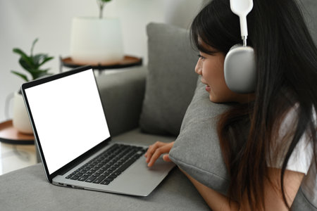 Happy young woman wearing headphone lying on couch and using her laptop at home.の写真素材