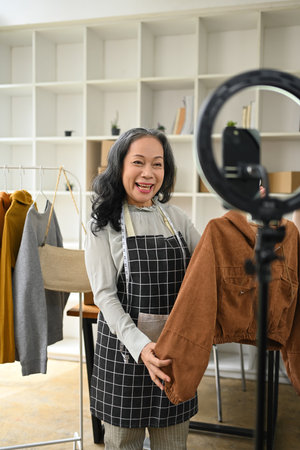 Cheerful senior lady small business owner selling clothes online by live streaming on mobile phone.の写真素材