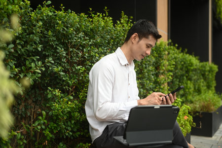 Millennial businessman using mobile phone while sitting outdoors cafe surrounded by green plants.の写真素材