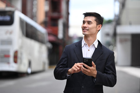 Confidence asian businessman holding smartphone looking away while walking on city street.の写真素材