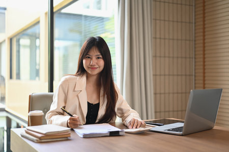Happy young businesswoman drinking coffee and reading email on laptop at her office.の写真素材