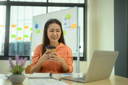 Attractive businesswoman holding paper cup of coffee while working with laptop in the office.の写真素材