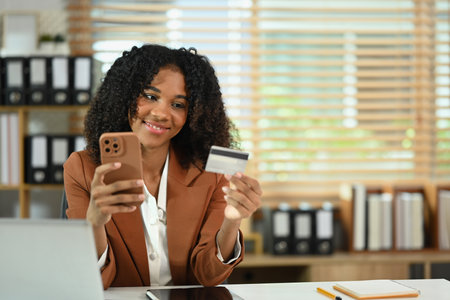 Smiling African American female worker holding mobile phone and credit card for financial transaction.の写真素材