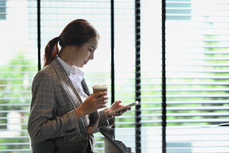 Portrait of Asian businesswoman drinking hot beverage and using mobile phone near office window.の写真素材