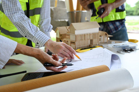 Construction engineers hard hats discussing plans over a table with blueprints, laptop, and...
