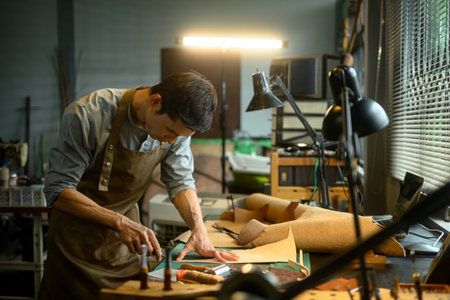 A craftsman working diligently, cutting leather in his workshop with various tools.の写真素材