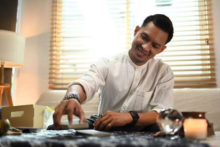 Smiling male fortune teller picking tarot card at table with burning candles and crystal ball. Forecasting and divination concept.の写真素材