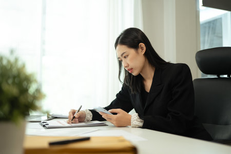 Focused businesswoman holding mobile phone and writing on clipboard.の写真素材