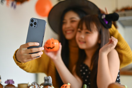 Happy smiling mother taking a picture of cute daughter with mobile phone decorating spooky cupcakes in kitchen.の写真素材