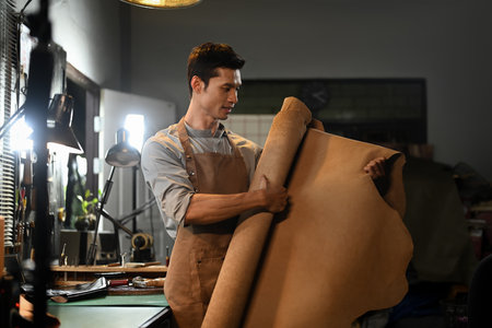 Craftsman working with large roll of leather in his workshop surrounded by tools and materials.