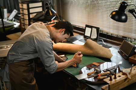 Concentrated male artisan cutting leather parts at table in his workshop.