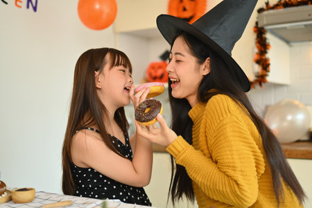 Cute little girl and mother eating sweet donut in kitchen. Halloween celebration concept.