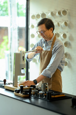 Senior male barista wearing an apron preparing a fresh cup of coffee using a modern espresso...