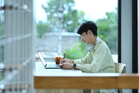 Concentrated young asian man writing in a notebook while working remotely with laptop at modern...
