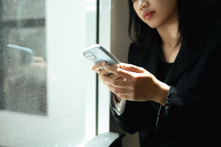 Pensive young businesswoman sitting near window, typing message on her mobile phone.