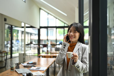 Charming young businesswoman having pleased phone conversation while drinking coffee by window.の写真素材