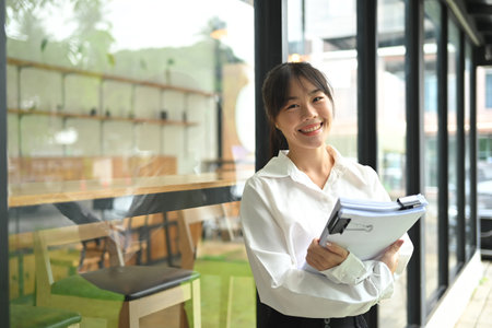 Smiling Asian female employee holding documents leaning against mirrored wall of office building.の写真素材