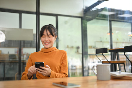 Portrait of smiling Asian woman freelancer sitting on coffee shop and checking her mobile phone.の写真素材