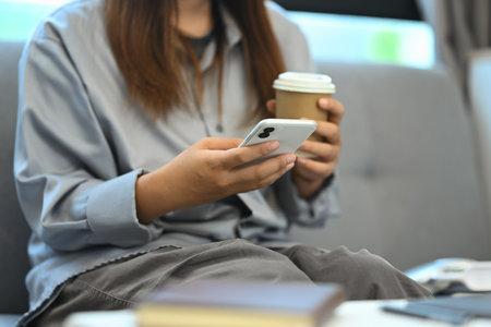 Young woman holding a paper cup of coffee and checking social media on her smartphone.の写真素材