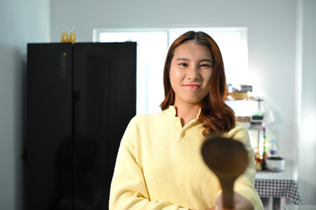 Playful young asian woman holding a spatula standing in the kitchen. Domestic life and cooking concept.の写真素材