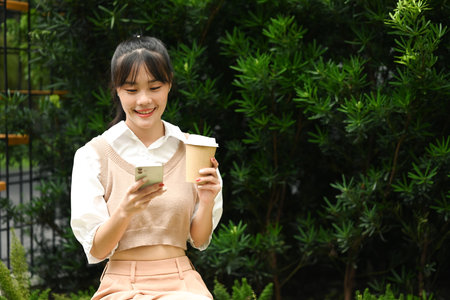 Smiling young asian woman holding paper cup of coffee and using mobile phone outdoor.