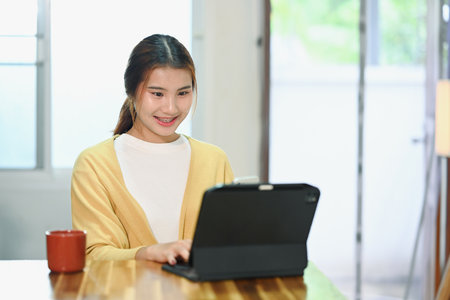 Cheerful young woman having virtual meeting or online study session on digital tablet, sitting in the kitchen.の写真素材
