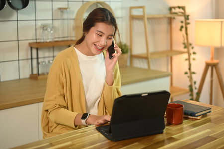 Cheerful young female freelancer talking on mobile phone and using digital tablet at a kitchen counter.の写真素材