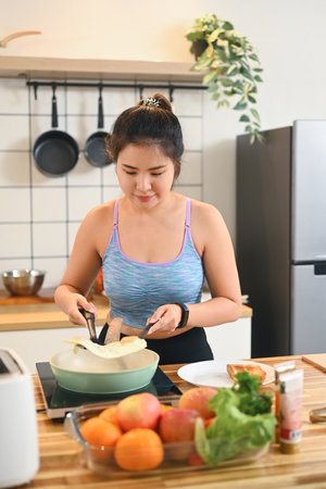 Young asian woman in sports wear preparing healthy breakfast, frying eggs in a kitchen.の写真素材