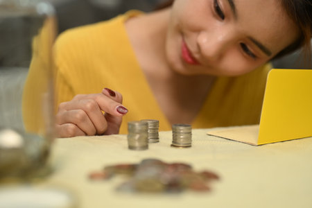 Cheerful young woman counting and stacking coins on a table. Saving money and financial responsibility concept.の写真素材