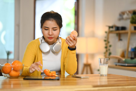 Pretty young woman sitting at kitchen table with a glass of milk, eating an orange and using a digital tablet.の写真素材