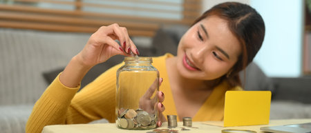 Happy young woman putting coins into a glass jar. Financial planning, budgeting or saving money...