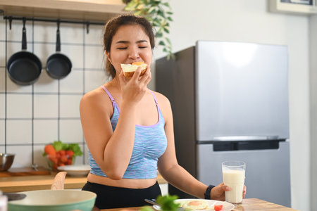 Young woman eating a sandwich and drinking milk, enjoying her meal in a cozy kitchen.の写真素材
