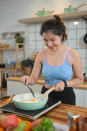 Beautiful young sportswoman frying eggs for breakfast in kitchen.の写真素材