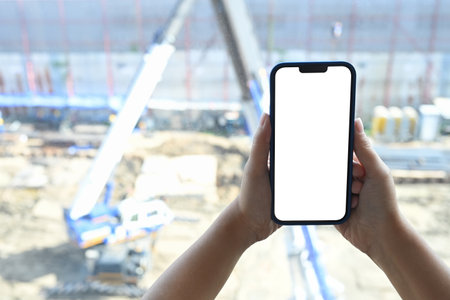Close up of hands holding smartphone with white empty screen on blurred construction site...