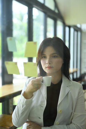 Thoughtful businesswoman holding drinking coffee and reading adhesive note on a glass wall in office.の写真素材