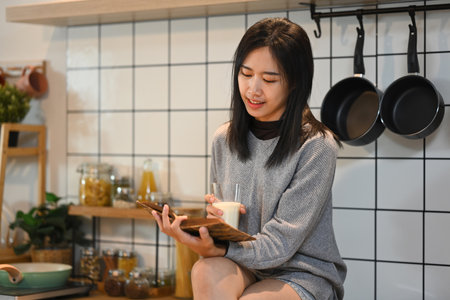 Happy young woman drinking milk and reading book while sitting on kitchen counter.の写真素材
